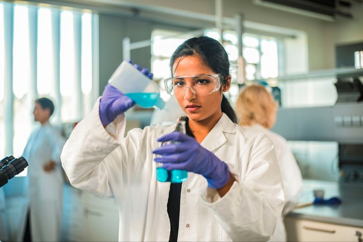 A young female scientist pours blue liquid into beaker. She is wearing a white lab coat.