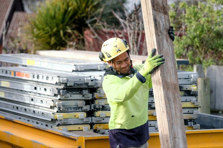 A male construction worker carries wooden plank. Behind him is a trailer of metal beams.