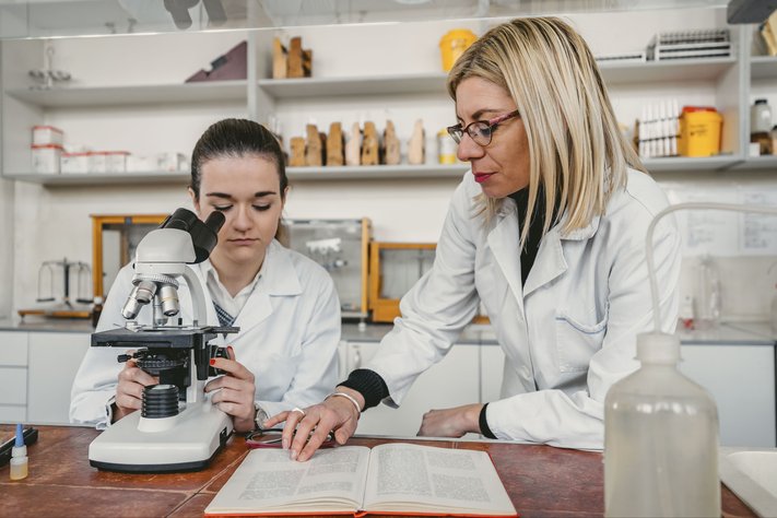 Female pharmacist guides female trainee pharmacist. They are reading a book and using a microscope.