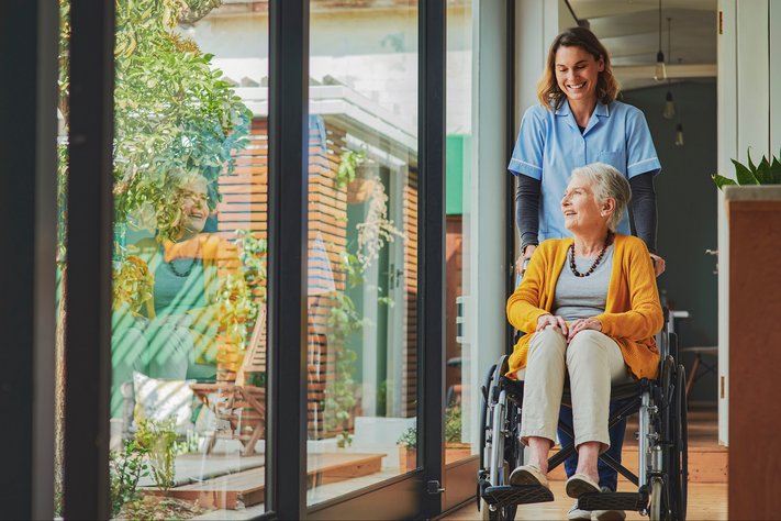 A female nurse pushes senior female patient in wheelchair through room.