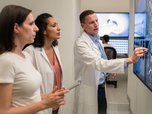 Three medical professional observe an x-ray scan of a brain. A male radiologist points to the image on the screen. They are wearing white lab coats.