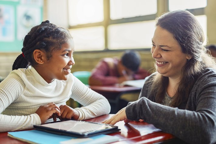 A female school teacher talks to female student at her desk.
