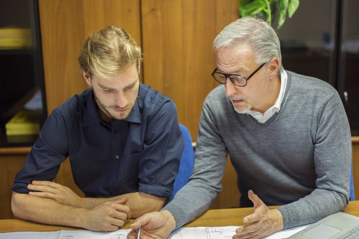 An old male accountant guides younger male accountant in an office.