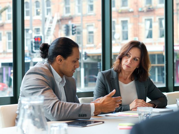 A man and woman sit a table in a boardroom in discussion. The male is gesturing in explanation and the female looks at him as she listens. There is paper, pens and an iPad in front of them.