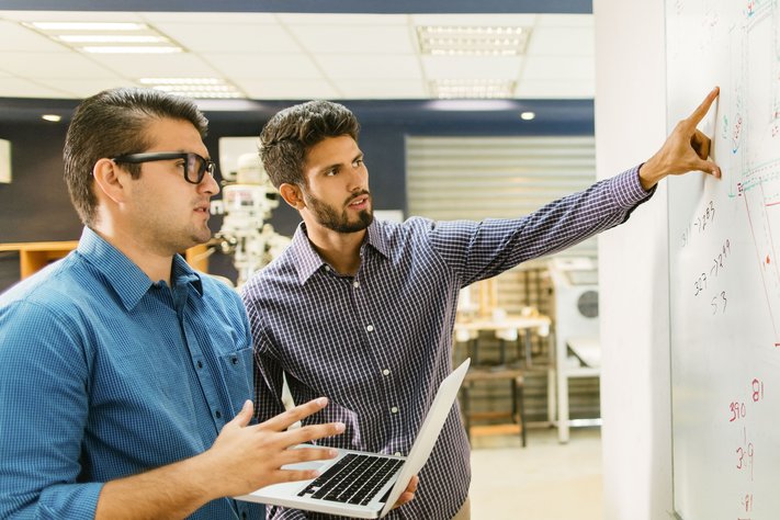 Two male professionals discuss project using a workspace whiteboard.