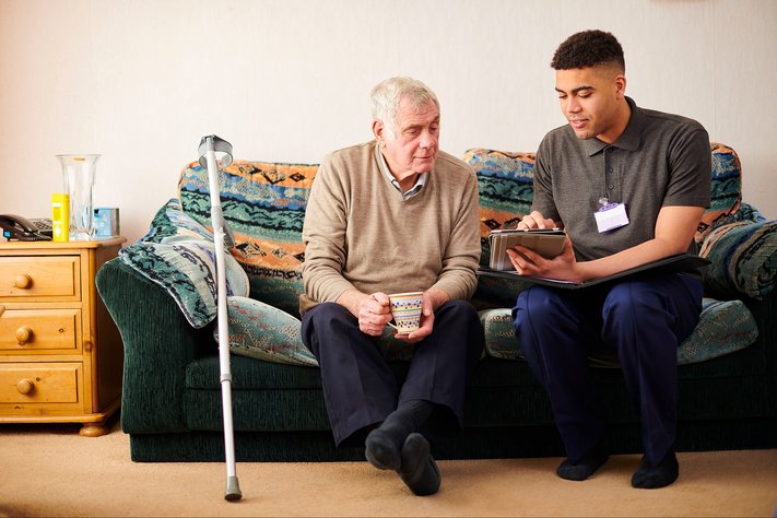 A male support worker sits down with elderly man on the couch. One crutch leans on the couch.