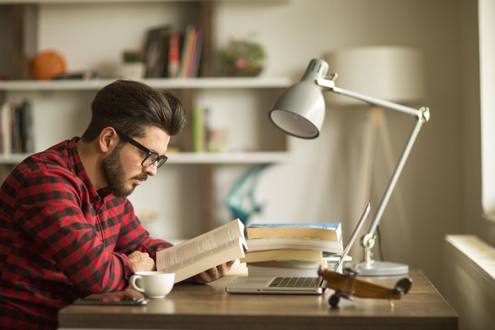 A young man reads a book at his office desk.