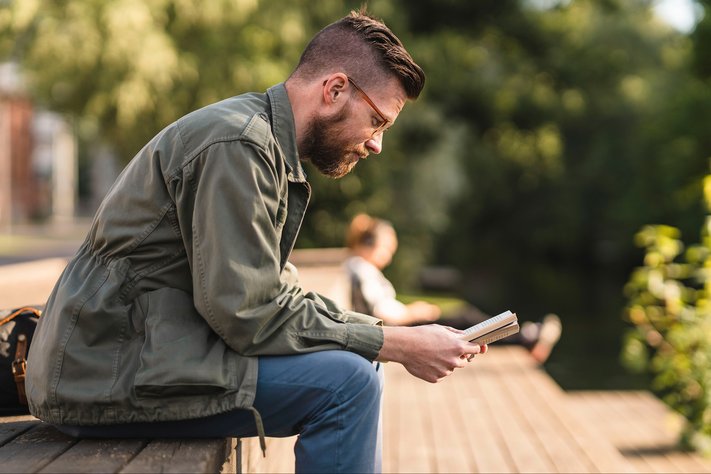 A man sits down to read a book in a sunny outdoor setting.