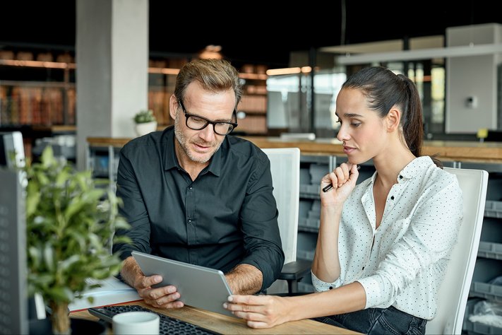 Male businessman shows younger female businesswoman computer in modern workspace.