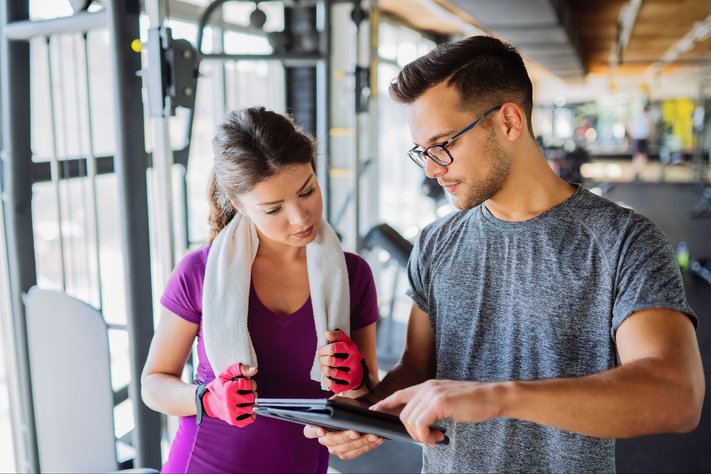 Male personal trainer shows computer tablet to female client. They are in a gym.