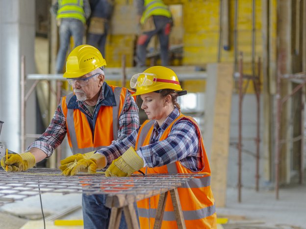 A man and woman in orange and yellow protective clothing work at a construction site measuring steel mesh before cutting it.