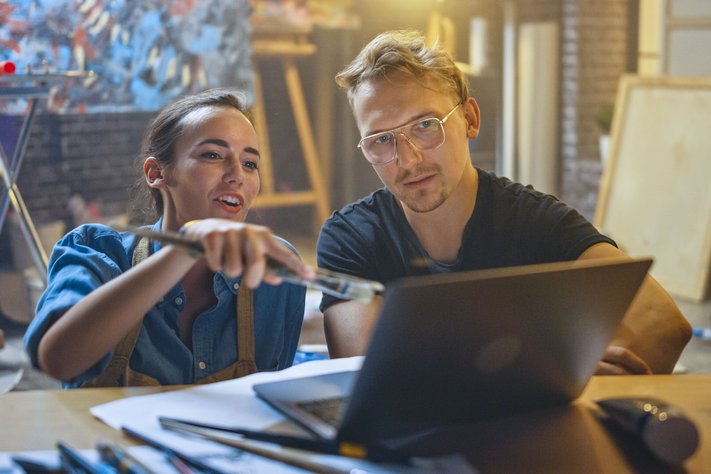 Two artists sit in their studio viewing a laptop. Behind them are oil paintings and tools.