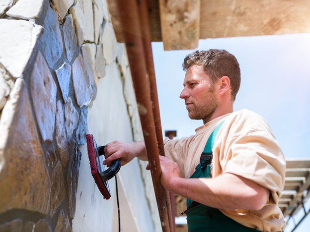 A man wearing a beige shirt and green overalls spreads concrete stone on a brick wall outdoors.