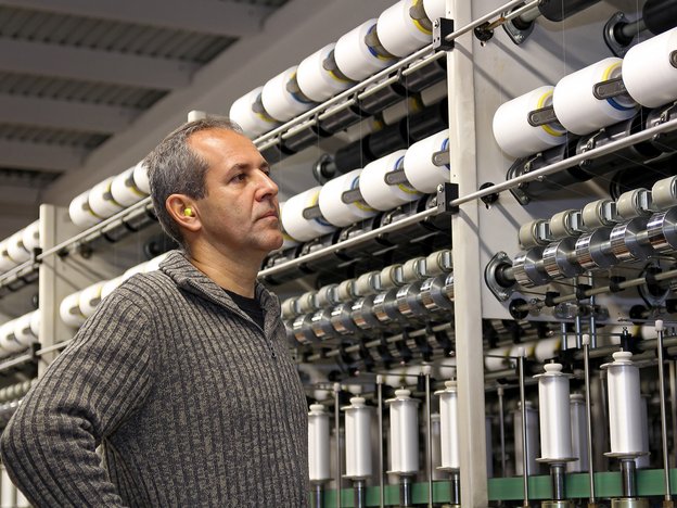 A man wearing a grey jersey with ear plugs works in a textiles factory.