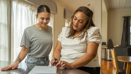 A mother and daughter discuss handwritten notes in a notebook. They stand at a table in their living room.