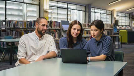 Teacher sits with two teenagers in library.