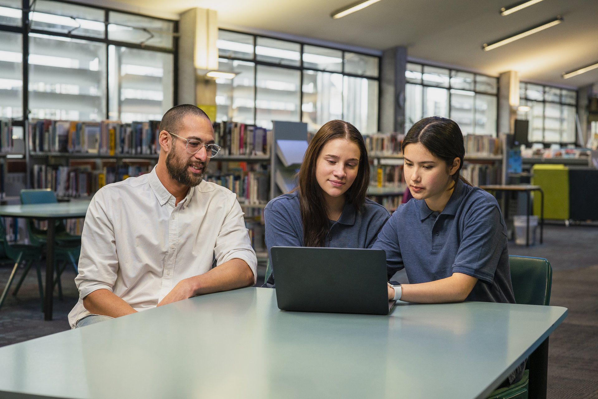 Teacher sits with two teenagers in library.