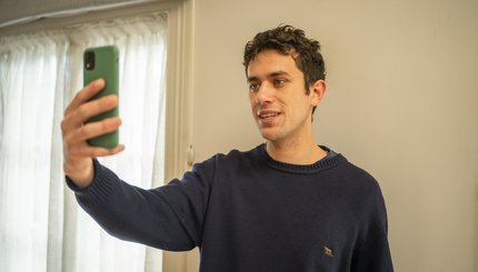 A young man holds up his mobile phone. He videos himself in his home.