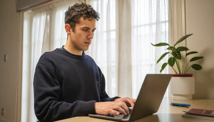 Young man seated at a desk in a house typing on a laptop.