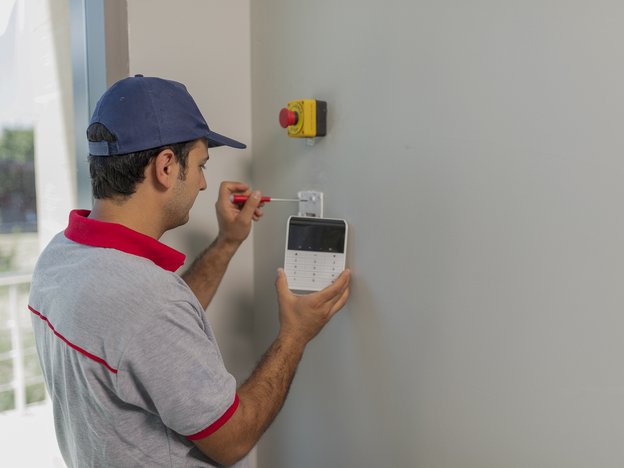 A man fixes an alarm on a white wall using a screwdriver. He wears a blue cap.