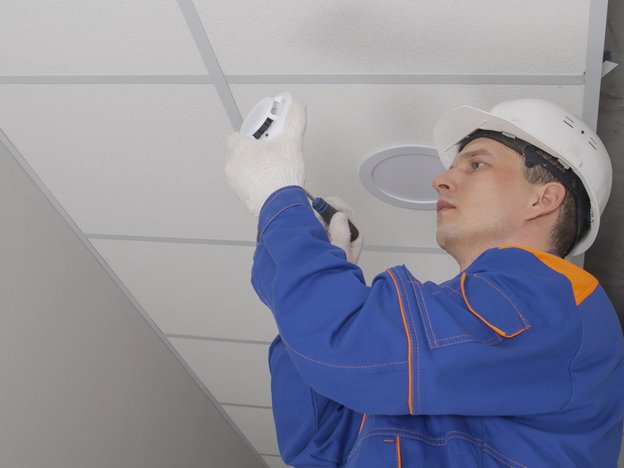 A man fixes an alarm on a ceiling using a screwdriver. He wears blue overalls, white gloves and a white helmet.