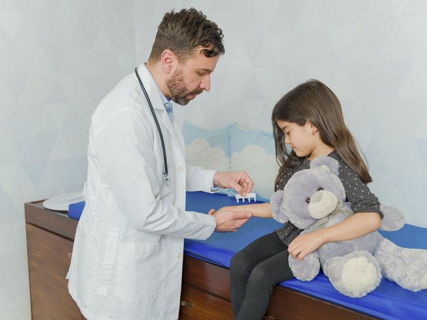 A man does a prick test on a child's arm. The man wears a lab coat and the girl sits on a medical bed. She holds a toy bear.