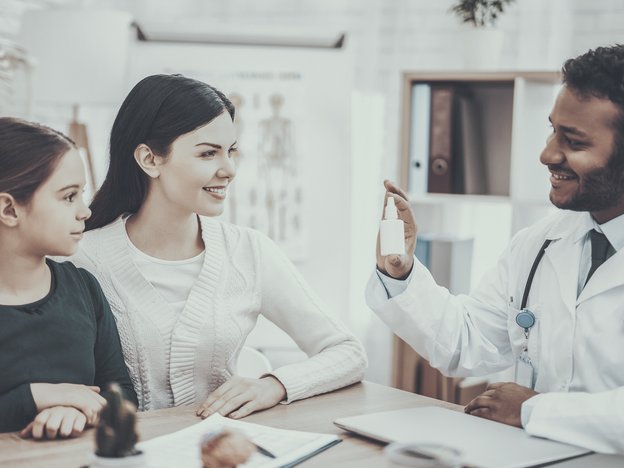 A man displays a small bottle to a woman and a girl. The man wears a lab coat and a stethoscope.