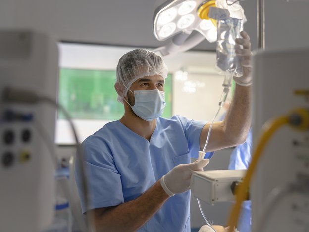 A male medical professional checks an IV bag. He is in an operating room wearing blue scrubs a hair net and a mask.
