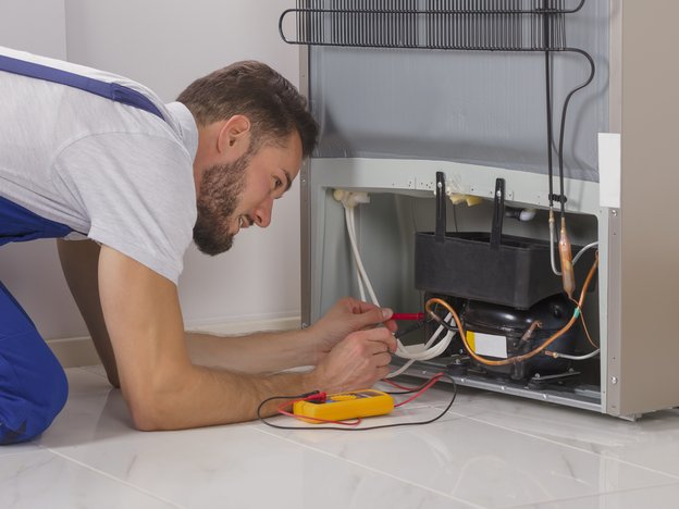 A man kneels on the ground to fix the wiring of an appliance using a screwdriver. He wears blue overalls.