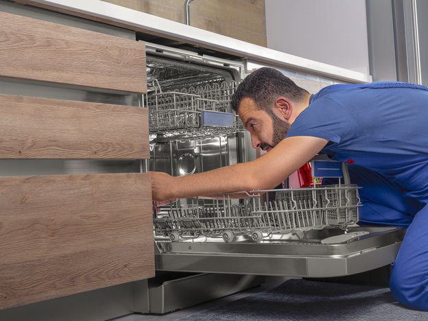 A man kneels on the ground to inspect a washing machine in a kitchen. He wears blue overalls.