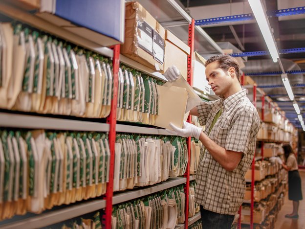A young man reads a document from shelving in an archival library. He is wearing white gloves.