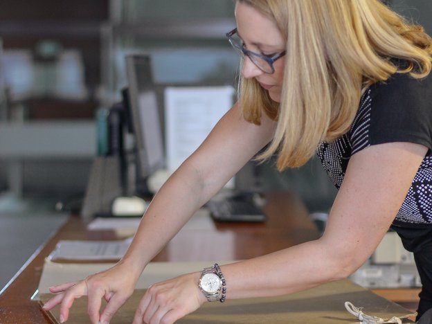 A woman ties a knot using string to hold an old paper folder together. She is surrounded by other archival documents on a bench.