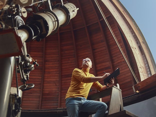 A man sits inside a stellar spectrum. He writes on a clipboard as he looks a spectroscope.