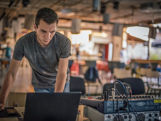 A man leans over a desk and looks at an open laptop. There is sound equipment on the desk.