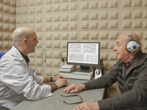 A man in a white lab coat tests the hearing of a man sitting opposite him who wears headphones and a brown jersey.
