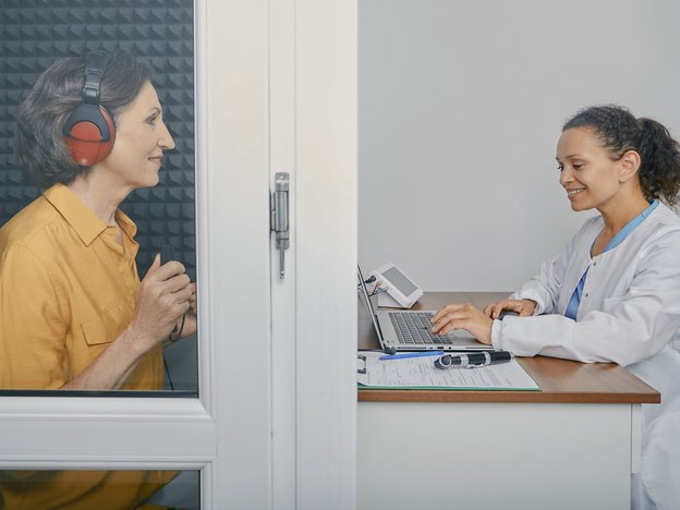 A woman in a white coat works on a laptop at a desk. A smiling lady sits in a cubicle opposite her with headphones on.
