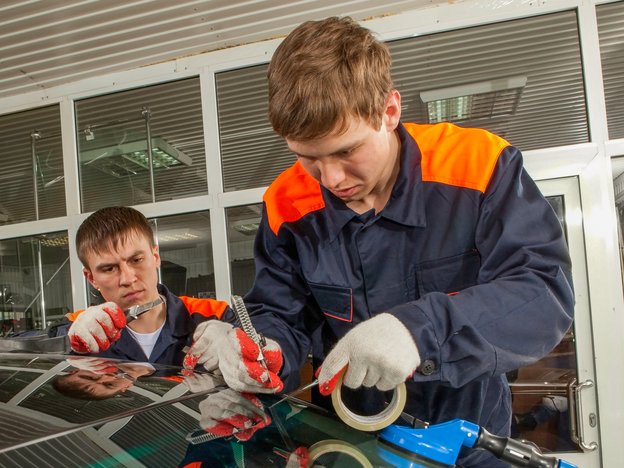 Two mechanics change a broken windshield in a workshop. They are wearing gloves and fluorescent orange and navy overalls.