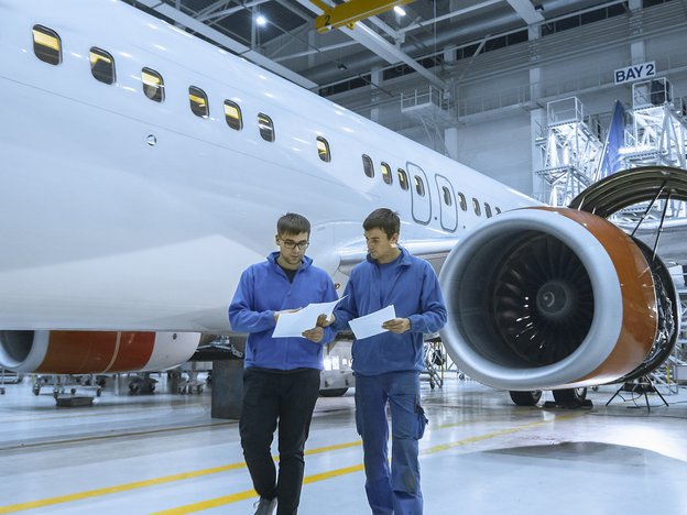 Two men stand in front of an airplane. They hold pieces of paper. They are in an aviation centre.