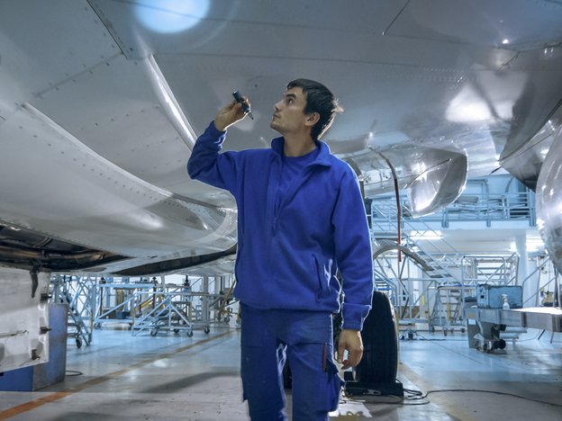 A man in a blue uniform stands under an airplane. He inspects under the carriage with a small torch.