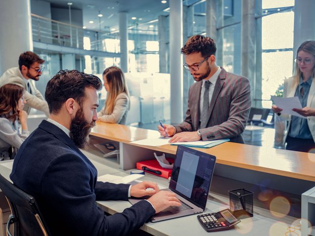 A man fills out a paper form at a front desk in a bank reception area. The bank teller is typing on computer and using a calculator. Several people queue up around them.