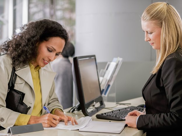 A woman fills out a paper form at a front desk with a pen, while a female receptionist stands behind the desk observing her.
