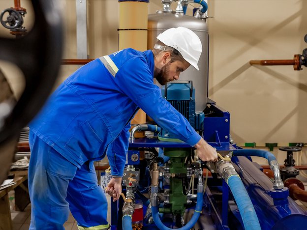 A male in a blue suit and white helmet fixes pipes in a boiler room.