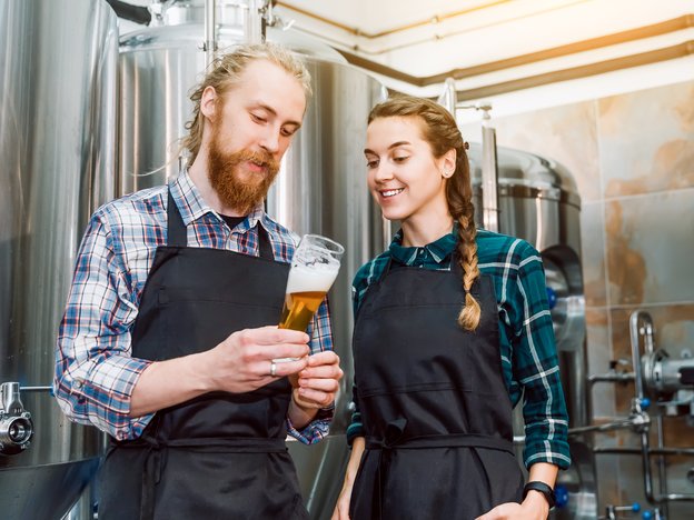 Two brewers inspect a glass of beer poured from a stainless steel tank.