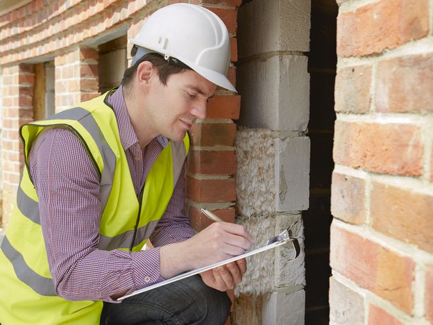A man kneels against a brick wall holding a clipboard and pen. He wears a white helmet and fluorescent yellow safety vest.