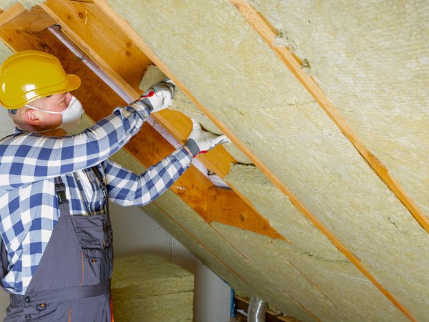 A builder places or removes insulation from the ceiling of an attic. They are wearing grey overalls and a yellow helmet.