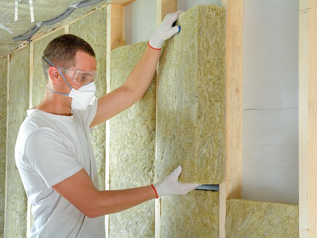 A man installs insulation within a wall. He is wearing safety glasses, a mask and gloves.