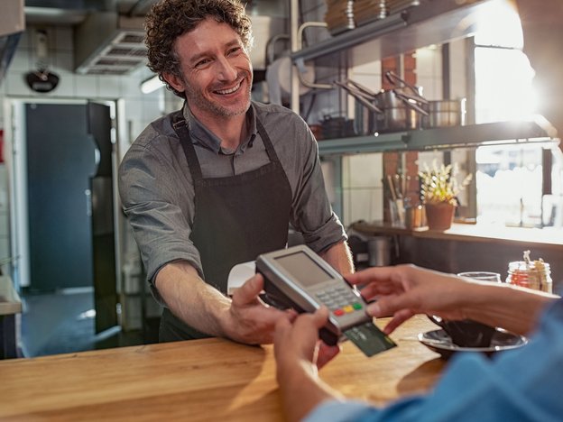A smiling man hands over an eftpos machine to a customer over the cash register. The customer is inserting his eftpos card and typing in his pine. They are in a cafe.