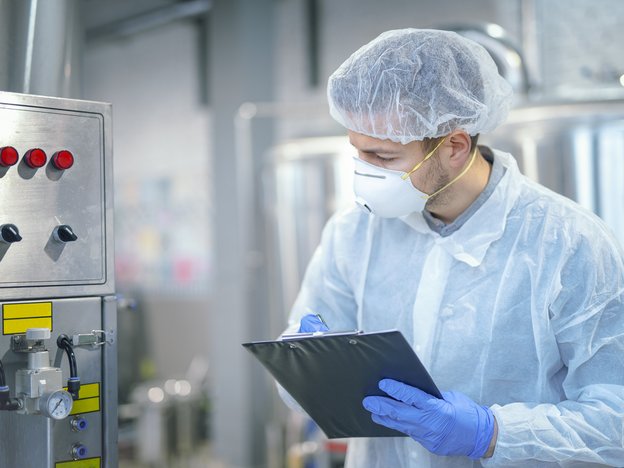 A man looks at a chemical machine and writes notes on a clipboard. He wears a protective fast mask, coat and a hair net