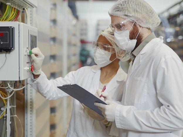 Two adults in white suits, white gloves and hairnets analyse a chemical plant box in a factory.