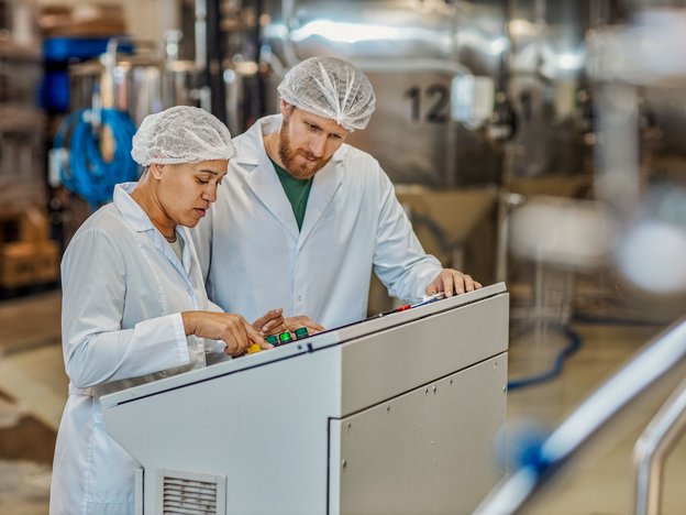 Two adults stand working on machine in a chemical plant operation factory. They woman points at a dial on the machine. They are wearing white coats and hairnets.